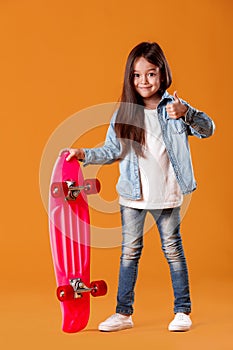 Stylish little child girl with skateboard in denim on orange background