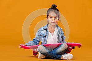 Stylish little child girl with skateboard in denim on orange background