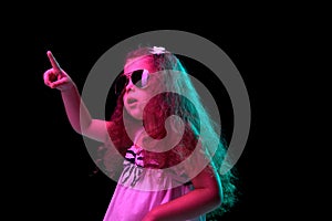 Stylish little girl, beginner fashion model wearing sunglasses posing isolated over dark background in neon light