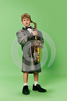 Stylish little school age boy in huge oversize retro style clothes and big male shoes posing with ukulele guitar