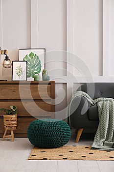 Stylish living room interior with knitted pouf, sofa and wooden chest of drawers