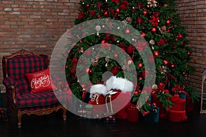 Stylish loft interior of room with Christmas fir tree and checkered armchair. Red sack santa claus with gifts near big Christmas f