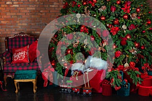 Stylish loft interior of room with Christmas fir tree and checkered armchair. Red sack santa claus with gifts near big Christmas f