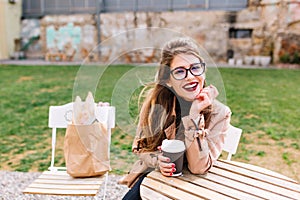 Stylish long-haired girl wearing brown coat and glasses, drinks latte in cafe after shopping with bags on chair behind