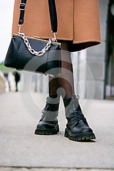 Stylish look. Close-up of a black handbag in the hands. Boots are black. Fashionable girl on the street.Black small handbag. Beige