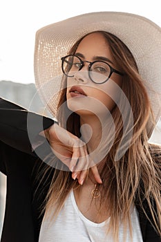 Stylish lovely young woman model in fashion glasses in fashionable straw hat in trendy black elegant blazer poses on street. Fresh