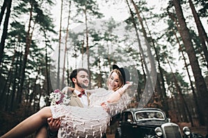 Stylish Loving wedding couple kissing and hugging in a pine forest near retro car