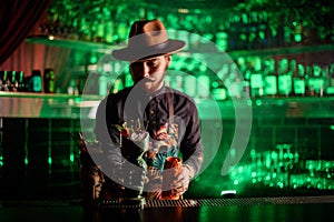 stylish male bartender in hat puts fancy glasses with ice cocktail on the bar counter