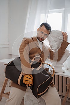 Stylish male embodies the old-money look, posing with a bag and tennis racket