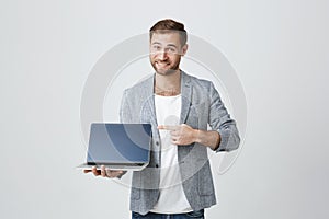 Stylish male enterpreneur with thick beard and trendy hairstyle posing against gray studio background with laptop in