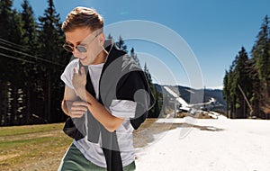 Stylish male tourist in casual clothes and sunglasses walks around the mountain resort with a smile on his face on a background of