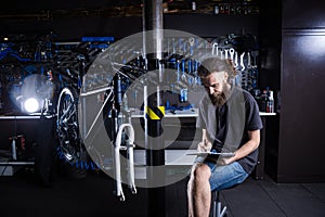 Stylish man with beard, cycle mechanic takes notes, checklist before repairing and servicing bike in bicycle shop workshop.