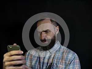 Stylish man with a beard and mustache looks at a mobile phone with a serious facial expression on a black background