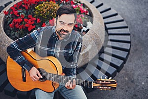 Stylish man with beard in a plaid shirt and jeans in the street near a park with flowers and a guitar in his hands. Macho with a b