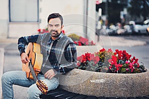 Stylish man with beard in a plaid shirt and jeans in the street near a park with flowers and a guitar in his hands. Macho with a b