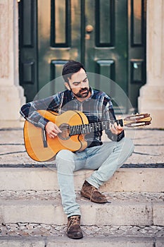 Stylish man with a beard in a plaid shirt and jeans in the street on the steps near the door. Macho with a beautiful smile sits in