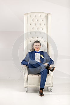 Stylish man in blue suit posing in white armchair at studio