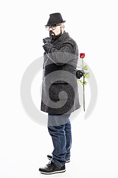 Stylish man in a coat and hat holds a red rose behind his back. White background. Vertical