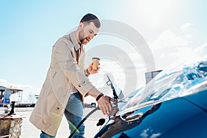 Stylish man with coffe cup in hand inserts plug into the electric car charging socket.