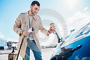 Stylish man with coffe cup in hand inserts plug into the electric car charging socket.