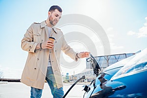 Stylish man with coffe cup in hand inserts plug into the electric car charging socket.