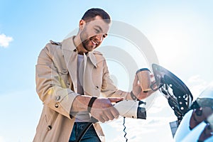 Stylish man with coffe cup in hand inserts plug into the electric car charging socket.