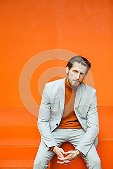 Stylish man in a fashionable suit sits against the background of an orange wall