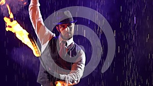 A stylish man in a hat rotates burning torches in his hands. The artist demonstrates his skills in the rain against a