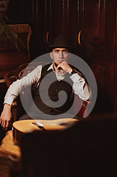 stylish man in hat is sitting in an armchair in a chic retro interior of restaurant