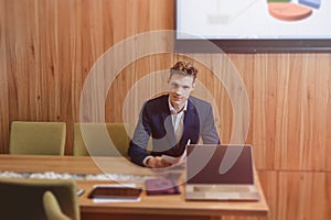 A stylish man in a jacket and shirt sits at the desk with his colleagues and works with documents at office