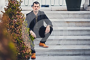A stylish man in a jacket, shirt, trousers and brown leather shoes sits on concrete steps. Portrait of a macho sitting on the stre