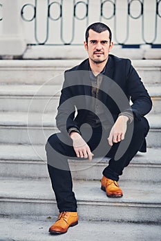 A stylish man in a jacket, shirt, trousers and brown leather shoes sits on concrete steps. Portrait of a macho sitting on the stre