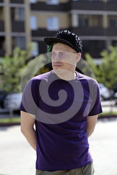 Stylish man in sunglasses standing on street. Side view of hipster with green dreadlocks in trendy outfit and sunglasses