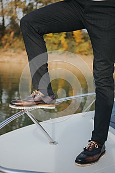 A stylish man in textured black suit pants and dark brogue shoes, standing on a boat