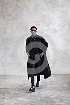 Stylish man walking in grey studio, wearing black coat, turtleneck, pants, loafers shoes. Fashion african american male