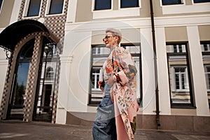 Stylish mature woman walks near city building, wearing cargo denim pants, pink floral trench coat, glasses. Urban street