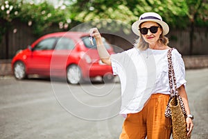 Stylish, mid-aged woman holding keys to new red car and smiling at camera. Ready for car trip.