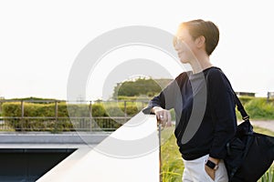 Stylish middle aged Asian beautiful woman standing in city park with green bushes, looking away with sunset behind