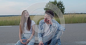 Stylish millennial teens sitting on skateboard on road and having romantic talk