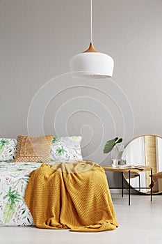 Stylish mirror and wooden bedside table next to big comfortable bed with leaf pattern on sheets and peach colored blanket, real