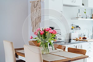 Stylish and modern boho, scandi interior of open space white kitchen with pink tulip flowers in vase on the wooden