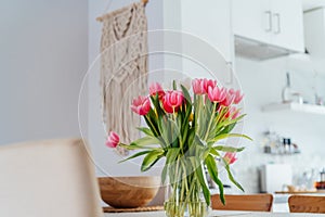 Stylish and modern boho, scandi interior of open space white kitchen with pink tulip flowers in vase on the wooden