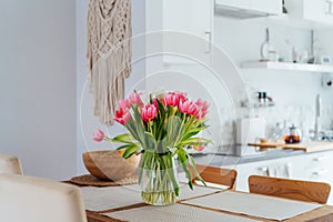 Stylish and modern boho, scandi interior of open space white kitchen with pink tulip flowers in vase on the wooden