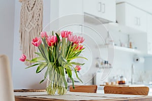 Stylish and modern boho, scandi interior of open space white kitchen with pink tulip flowers in vase on the wooden