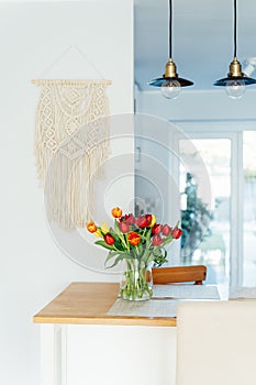 Stylish and modern boho, scandi interior of open space white kitchen with tulip flowers in vase on the wooden kitchen