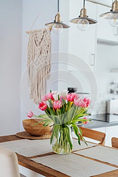 Stylish and modern boho, scandi interior of open space white kitchen with tulip flowers in vase on wooden kitchen