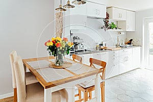 Stylish and modern boho, scandi interior of open space white kitchen with yellow and orange roses flower in vase on the