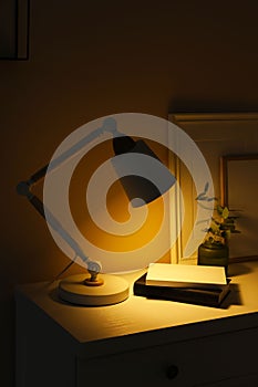 Stylish modern desk lamp, books and plant on white chest of drawers in dark room