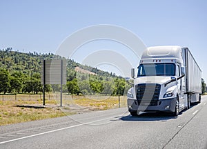 Stylish modern white big rig semi truck with dry van semi trailer transporting cargo running on the road along the orchards in