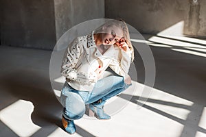 Stylish modern young blond woman in trendy jacket in blue ripped jeans in green fashionable cowboy boots posing sitting indoors